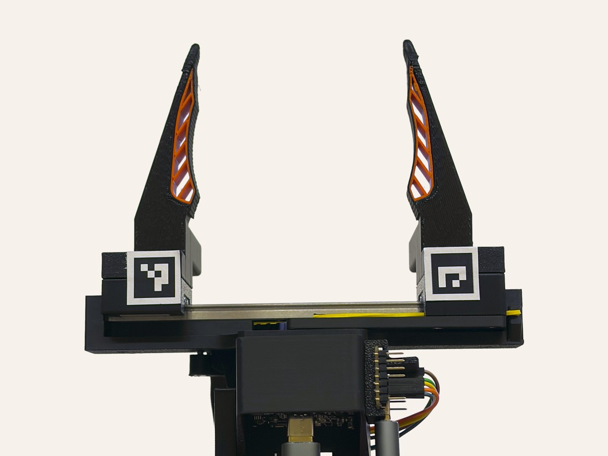 The UMI gripper with its jaws open in front of a white screen: one ArUco tag sits on each sledge, close to the camera.